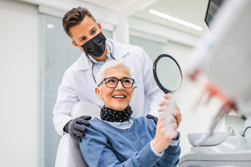 a picture of a periodontist wearing a mask who is talking to a patient about what can affect the cost of her dental implants as she smiles at herself through a mirror.