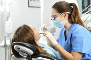 Dentist wearing eyeglasses gloves and mask examining a patient tooth with a dental probe and a mirror in a clinic box with equipment in the background.